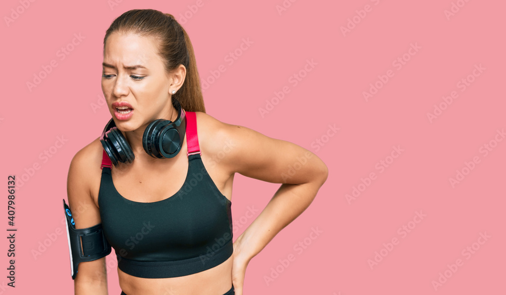 Beautiful blonde woman wearing gym clothes and using headphones suffering of backache, touching back with hand, muscular pain Beautiful blonde woman wearing gym clothes and using headphones suffering of backache, touching back with hand, muscular pain