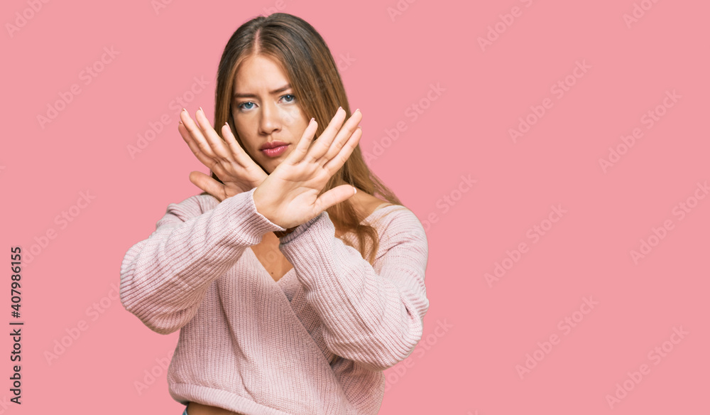 Beautiful blonde woman wearing casual winter pink sweater rejection expression crossing arms and palms doing negative sign, angry face Beautiful blonde woman wearing casual winter pink sweater rejection expression crossing arms and palms doing negative sign, angry face
