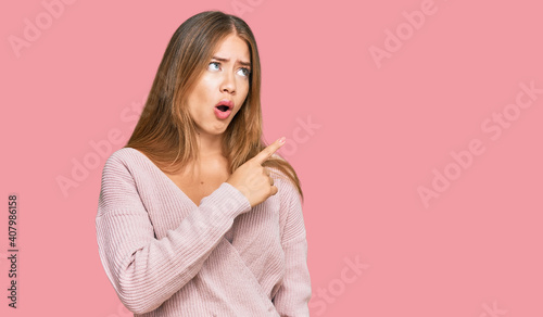 Beautiful blonde woman wearing casual winter pink sweater surprised pointing with finger to the side, open mouth amazed expression. © Krakenimages.com