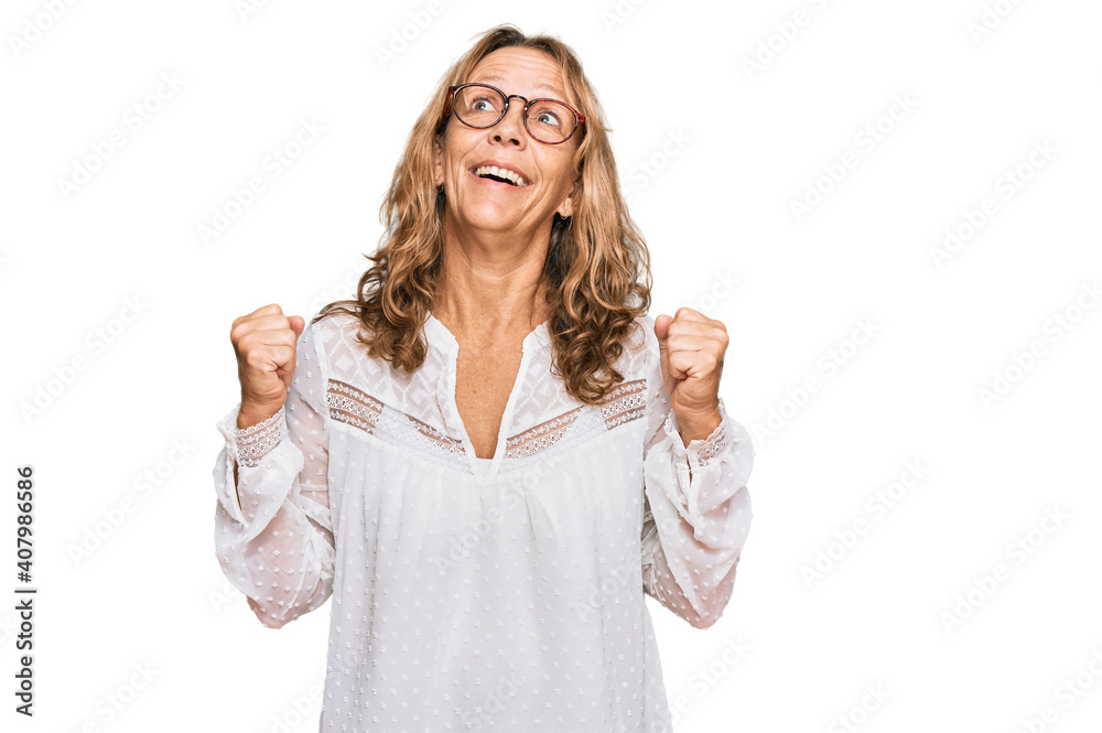 Middle age blonde woman wearing casual white shirt and glasses celebrating surprised and amazed for success with arms raised and open eyes. winner concept. Middle age blonde woman wearing casual white shirt and glasses celebrating surprised and amazed for success with arms raised and open eyes. winner concept.