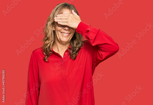 Middle age blonde woman wearing casual shirt over red background smiling and laughing with hand on face covering eyes for surprise. blind concept. © Krakenimages.com