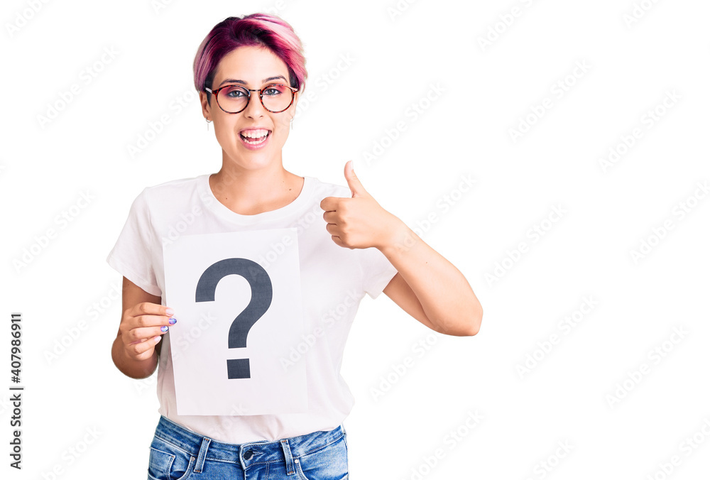 Young beautiful woman with pink hair holding question mark smiling happy and positive, thumb up doing excellent and approval sign Young beautiful woman with pink hair holding question mark smiling happy and positive, thumb up doing excellent and approval sign