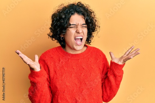 Young hispanic woman with curly hair wearing casual winter sweater celebrating mad and crazy for success with arms raised and closed eyes screaming excited. winner concept