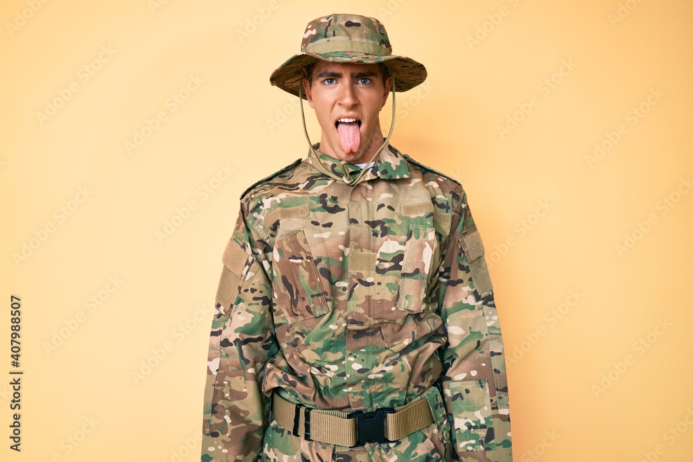 Young handsome man wearing camouflage army uniform sticking tongue out happy with funny expression. emotion concept.