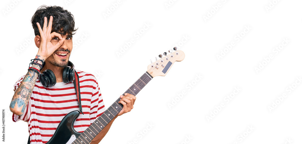 Young hispanic man playing electric guitar smiling happy doing ok sign with hand on eye looking through fingers