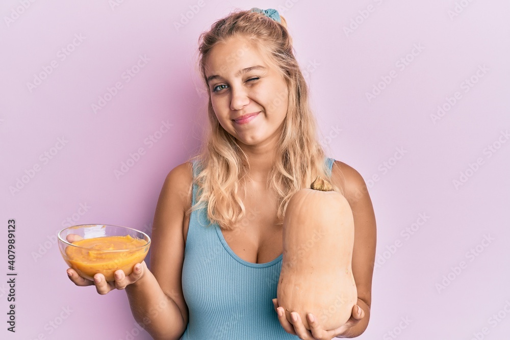 Young blonde girl holding fresh pumpkin and soup winking looking at the camera with sexy expression, cheerful and happy face. Young blonde girl holding fresh pumpkin and soup winking looking at the camera with sexy expression, cheerful and happy face.