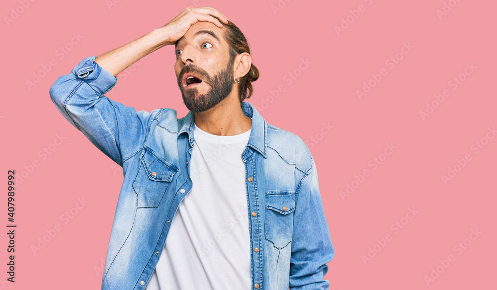 Fototapeta premium Attractive man with long hair and beard wearing casual denim jacket surprised with hand on head for mistake, remember error. forgot, bad memory concept.