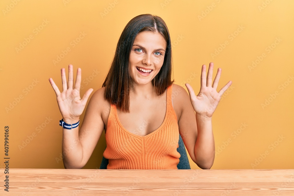 Young caucasian woman wearing casual clothes sitting on the table showing and pointing up with fingers number ten while smiling confident and happy. Young caucasian woman wearing casual clothes sitting on the table showing and pointing up with fingers number ten while smiling confident and happy.