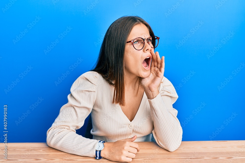 Young caucasian woman wearing casual clothes and glasses shouting and screaming loud to side with hand on mouth. communication concept. Young caucasian woman wearing casual clothes and glasses shouting and screaming loud to side with hand on mouth. communication concept.
