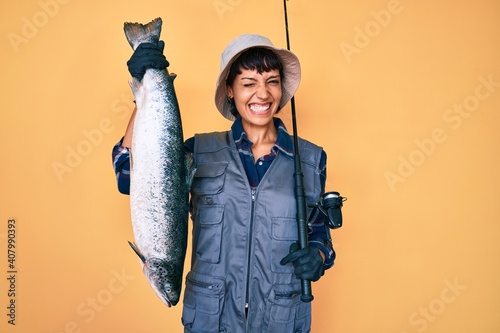 Fotografie Beautiful brunettte fisher woman holding fishing rod and raw salmon winking looking at the camera with sexy expression, cheerful and happy face
