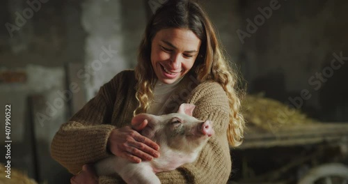 Cinematic shot of young female farmer is caressing with love and care ecologically grown newborn pink piglet used for biological genuine products industry in pigpen of countryside pigs breeding farm.