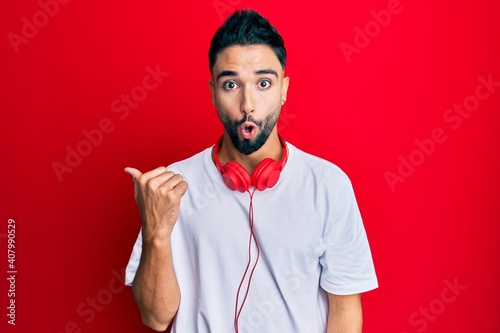 Young man with beard listening to music using headphones surprised pointing with hand finger to the side, open mouth amazed expression. © Krakenimages.com