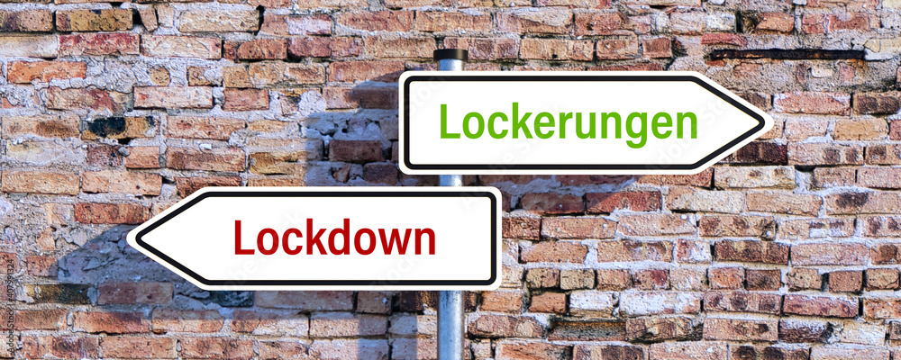 two street signs pointing in different directions showing the German message for LOCKDOWN and EASING in front of a brick wall two street signs pointing in different directions showing the German message for LOCKDOWN and EASING in front of a brick wall