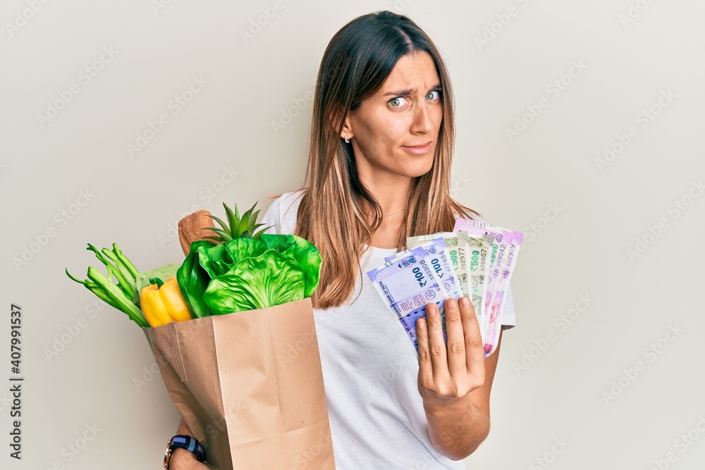 Brunette young woman holding groceries and indian rupee banknotes clueless and confused expression. doubt concept. Brunette young woman holding groceries and indian rupee banknotes clueless and confused expression. doubt concept.