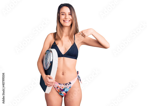 Foto Young beautiful girl wearing bikini holding weighing machine pointing finger to