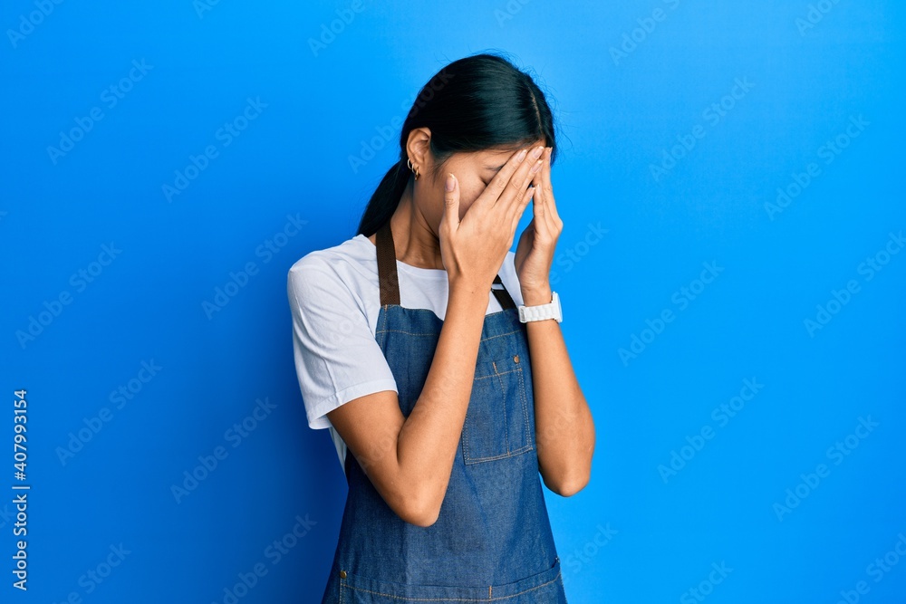 Young chinese woman wearing waiter apron with sad expression covering face with hands while crying. depression concept. Young chinese woman wearing waiter apron with sad expression covering face with hands while crying. depression concept.