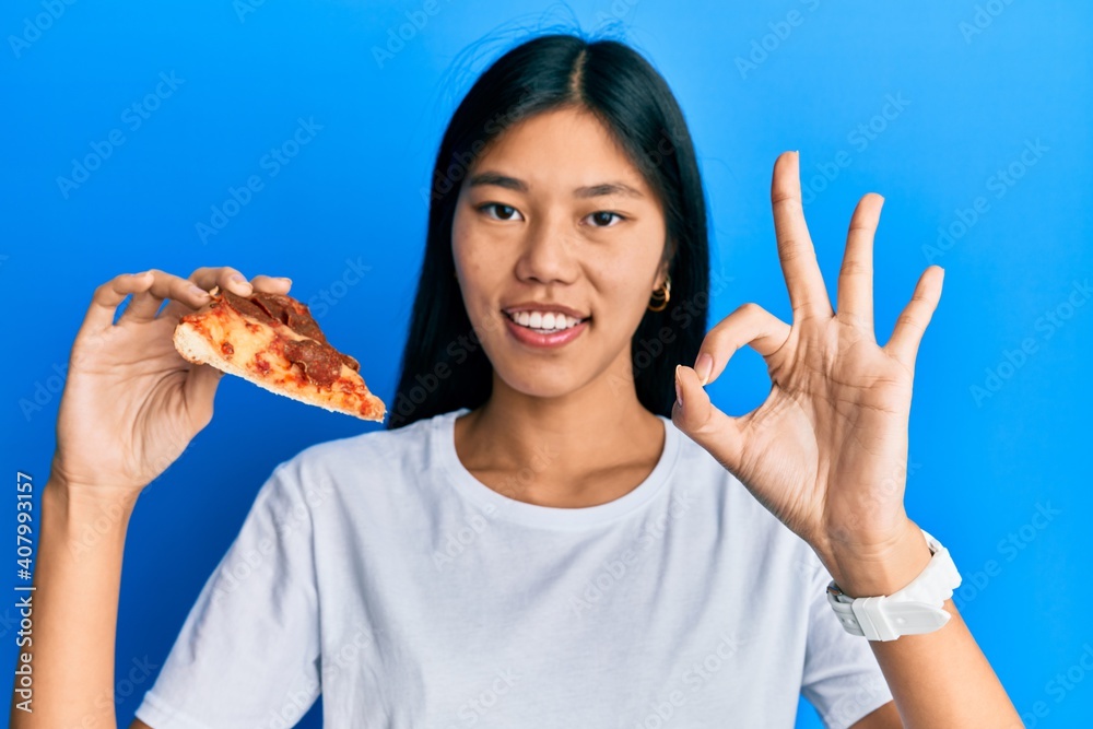 Young chinese woman eating tasty pepperoni pizza doing ok sign with fingers, smiling friendly gesturing excellent symbol Young chinese woman eating tasty pepperoni pizza doing ok sign with fingers, smiling friendly gesturing excellent symbol