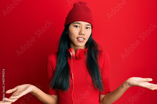 Young chinese woman listening to music using headphones clueless and confused expression with arms and hands raised. doubt concept.