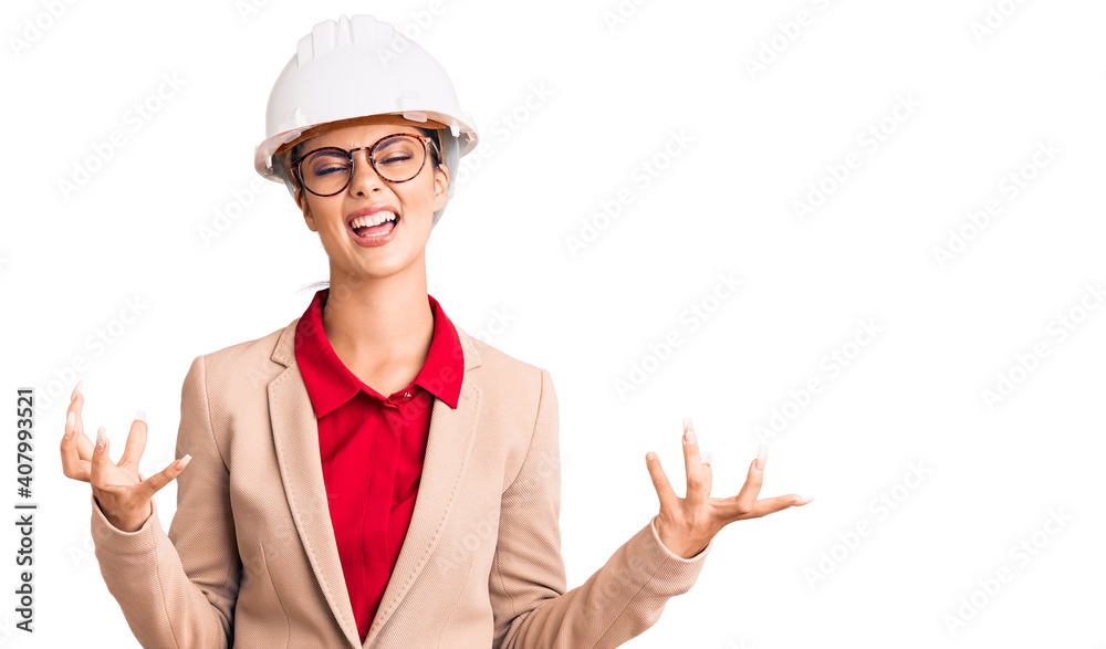 Young beautiful woman wearing architect hardhat crazy and mad shouting and yelling with aggressive expression and arms raised. frustration concept. Young beautiful woman wearing architect hardhat crazy and mad shouting and yelling with aggressive expression and arms raised. frustration concept.