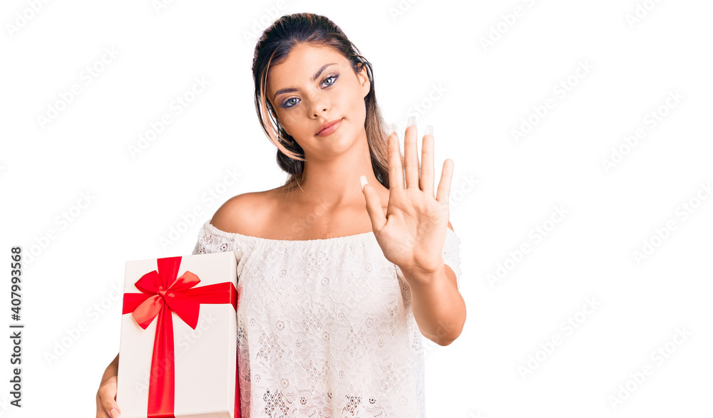Young beautiful woman holding gift with open hand doing stop sign with serious and confident expression, defense gesture Young beautiful woman holding gift with open hand doing stop sign with serious and confident expression, defense gesture