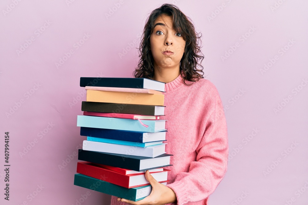 Young hispanic woman holding a pile of books puffing cheeks with funny face. mouth inflated with air, catching air. Young hispanic woman holding a pile of books puffing cheeks with funny face. mouth inflated with air, catching air.