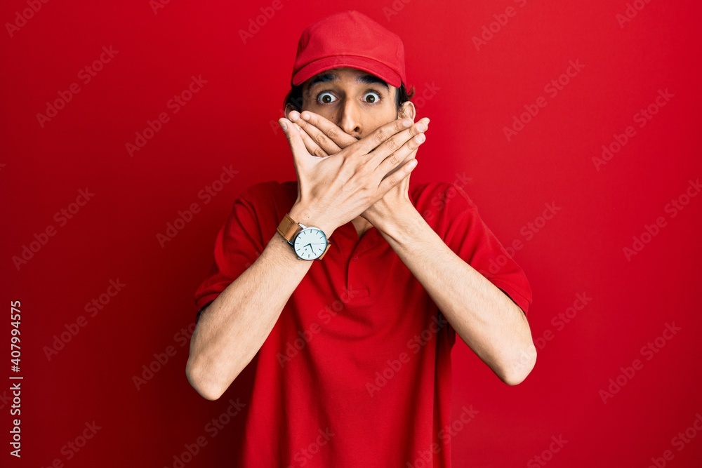 Young hispanic man wearing delivery uniform and cap shocked covering mouth with hands for mistake. secret concept. Young hispanic man wearing delivery uniform and cap shocked covering mouth with hands for mistake. secret concept.