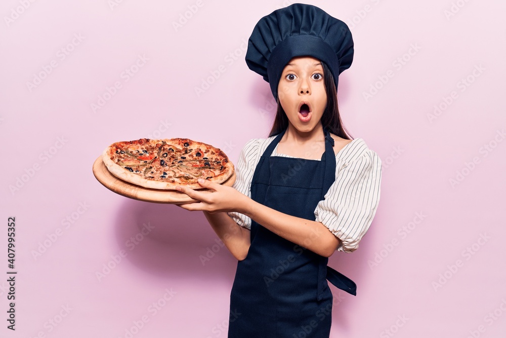 Beautiful child girl wearing cooker uniform holding italian pizza scared and amazed with open mouth for surprise, disbelief face Beautiful child girl wearing cooker uniform holding italian pizza scared and amazed with open mouth for surprise, disbelief face