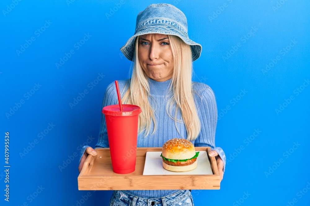 Young blonde woman eating a tasty classic burger and soda skeptic and nervous, frowning upset because of problem. negative person. Young blonde woman eating a tasty classic burger and soda skeptic and nervous, frowning upset because of problem. negative person.