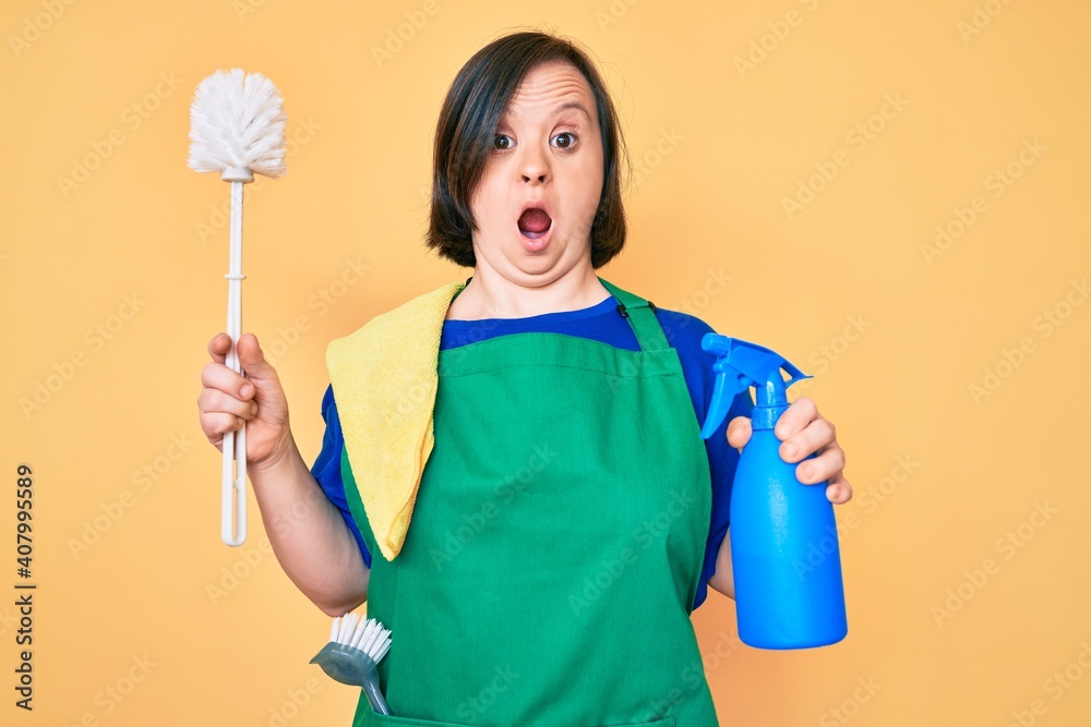 Brunette woman with down syndrome wearing apron holding scourer and toilet brush afraid and shocked with surprise and amazed expression, fear and excited face. Brunette woman with down syndrome wearing apron holding scourer and toilet brush afraid and shocked with surprise and amazed expression, fear and excited face.