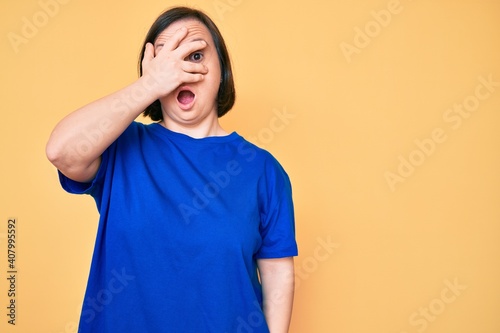 Brunette woman with down syndrome wearing casual clothes peeking in shock covering face and eyes with hand, looking through fingers afraid