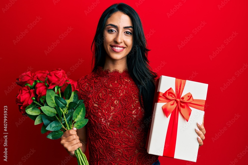 Beautiful hispanic woman holding anniversary gift and roses bouquet smiling with a happy and cool smile on face. showing teeth. Beautiful hispanic woman holding anniversary gift and roses bouquet smiling with a happy and cool smile on face. showing teeth.