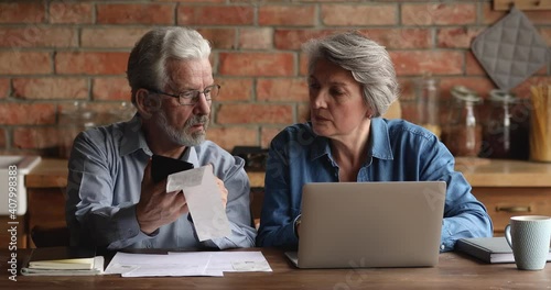 Mature spouses sit in kitchen using e-bank app on laptop, discussing analysing financial papers, cashiers checks, invoices, receipts of payment for services, family budget counting summary concept