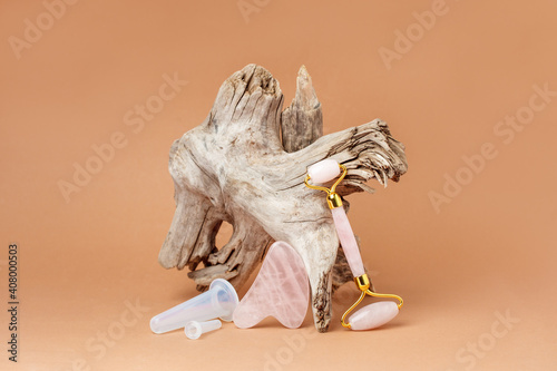 Fototapeta Naklejka Na Ścianę i Meble -  Modern luxury stylish beauty concept - Facial massage kit and wooden root on beige paper background. Face roller and gua sha massager made from natural pink stones, wellbeing, face fitness