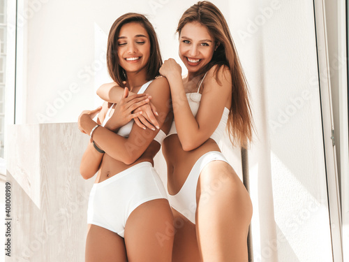 Portrait of two young beautiful smiling women in white lingerie. Sexy carefree models posing at the balcony in the early morning. Models hugging and enjoying their morning photo