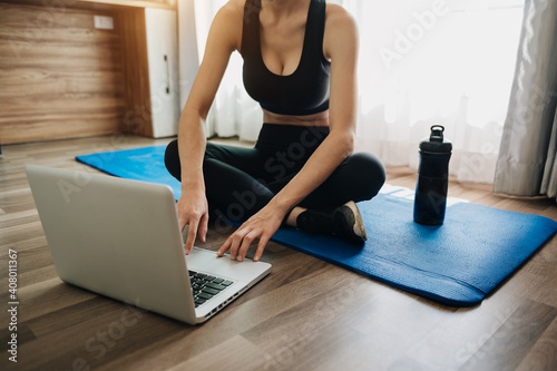  sporty woman in sportswear is sitting on the floor with dumbbells and a protein shake or a bottle of water and is using a laptop.