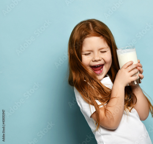 Cheerful happy small cute girl in white t-shirt standing with glass of milk yogurt kefir feeling excited with eyes closed over blue background, copy space. Happy childhood, healthy lifestyle concept photo