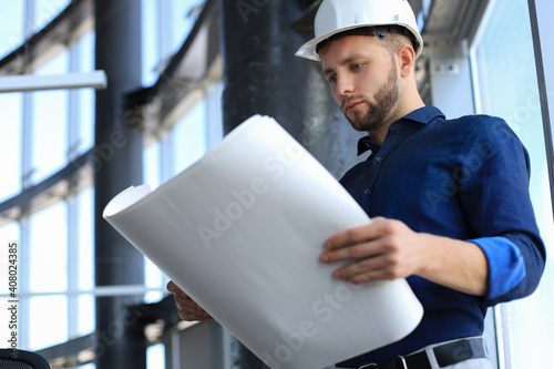 Confident young architect in navy shirt and hardhat holding a blueprint and looking at it.