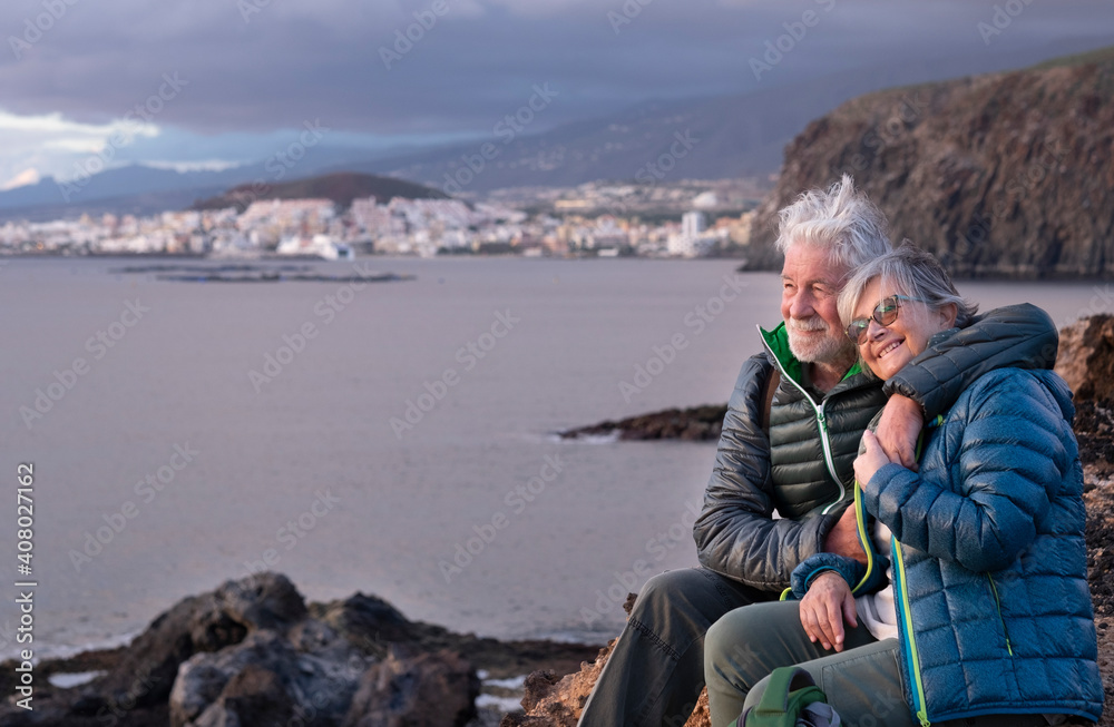 Fototapeta premium Happy white-haired senior couple hugs each other while sitting on the cliff on a winter day, enjoying the outdoors. Cloudy sky, coast and mountain in the background