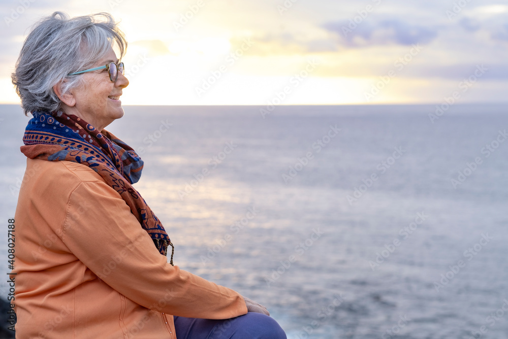 Relaxed senior grey-haired woman enjoying the freedom and the horizon over water. Sitting on the cliff in front to the sea in a winter cloudy day