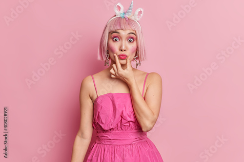 Pretty young Asian woman pouts lips dresses for costume party wears unicorn headband and fashionable dress has rosy hair professional makeup isolated over pink background. People style concept