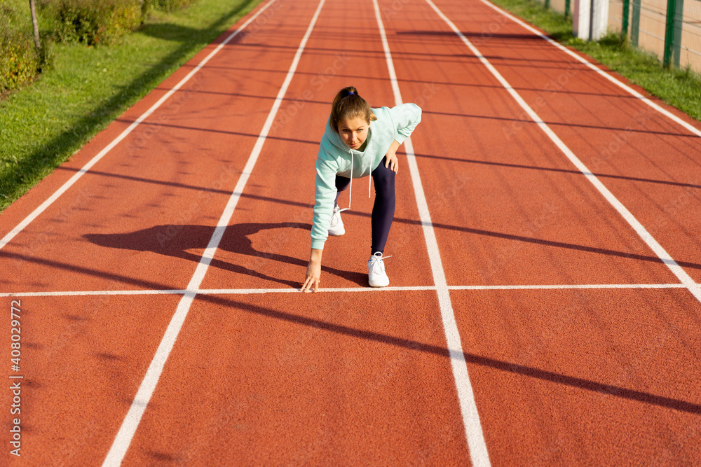 Training on the street, a young female athlete makes a workout on the street platform. Get ready, athlete in a low start on the treadmill, rubber stadium in the open air.