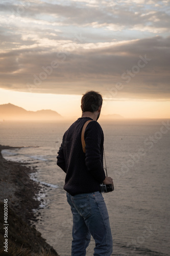 man from behind looking at the horizon in front of the sea on top of a cliff during sunset and with a camera on his shoulder  photo