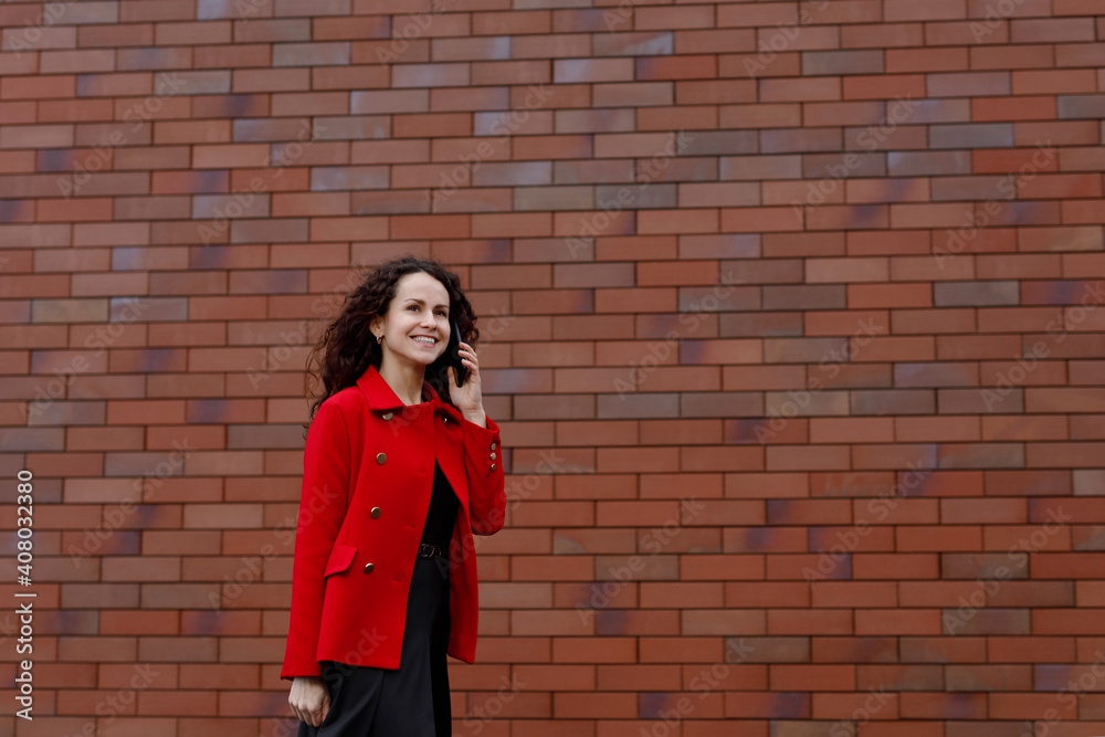 Smiling business woman in red, hair in wind, talking on the smartphone while walking outside, near brick wall. woman happy using mobile phone app for conversation. Space for text. Smiling business woman in red, hair in wind, talking on the smartphone while walking outside, near brick wall. woman happy using mobile phone app for conversation. Space for text.
