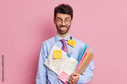 Indoor studio shot of male student makes funny grimace foolishes around crosses eyes and sticks out tongue prepares for exam in math holds papers and sticker notes on clothes isolated over pink wall