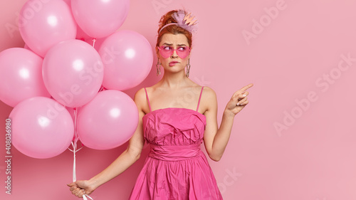 Puzzled redhead young woman dressed in glamour outfit wears everything pink poses on hens party with inflated balloons points at empty space bites lips shows place for your advertising content