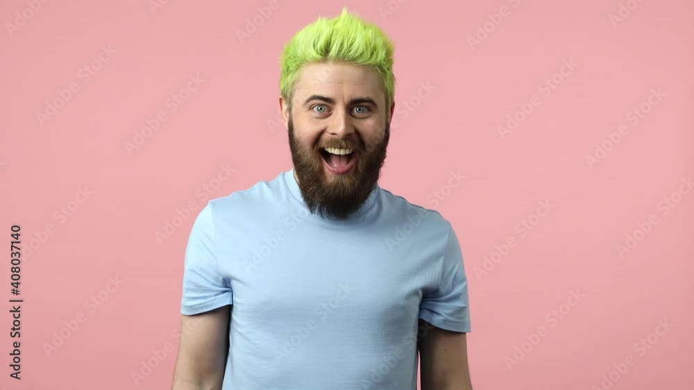 Extremely positive bearded man with dyed bright green hair suddenly appearing and waving hand looking at camera, greeting and welcoming you. Indoor studio shot isolated on pink background Extremely positive bearded man with dyed bright green hair suddenly appearing and waving hand looking at camera, greeting and welcoming you. Indoor studio shot isolated on pink background