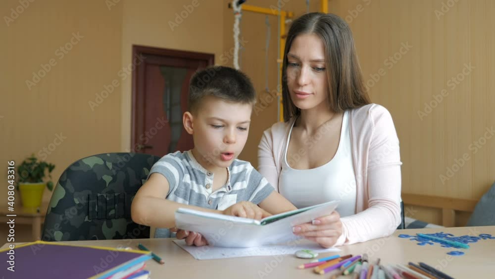 Young mother sitting at a table at home helping her son with his homework from school Young mother sitting at a table at home helping her son with his homework from school
