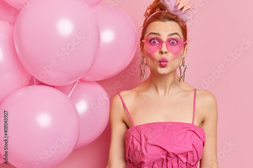 Surprised lovely young redhead woman makes lips in kiss wears trendy heart shaped sunglasses and dress comes on bachelorette party with inflated balloons stands indoor against pink background
