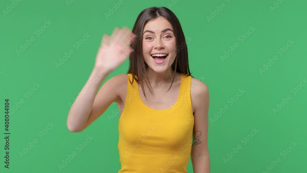 Excited cheerful brunette young woman 20s years old in yellow basic tank top posing isolated on green background in studio. People lifestyle concept. Waving and greeting with hand as notices someone Excited cheerful brunette young woman 20s years old in yellow basic tank top posing isolated on green background in studio. People lifestyle concept. Waving and greeting with hand as notices someone