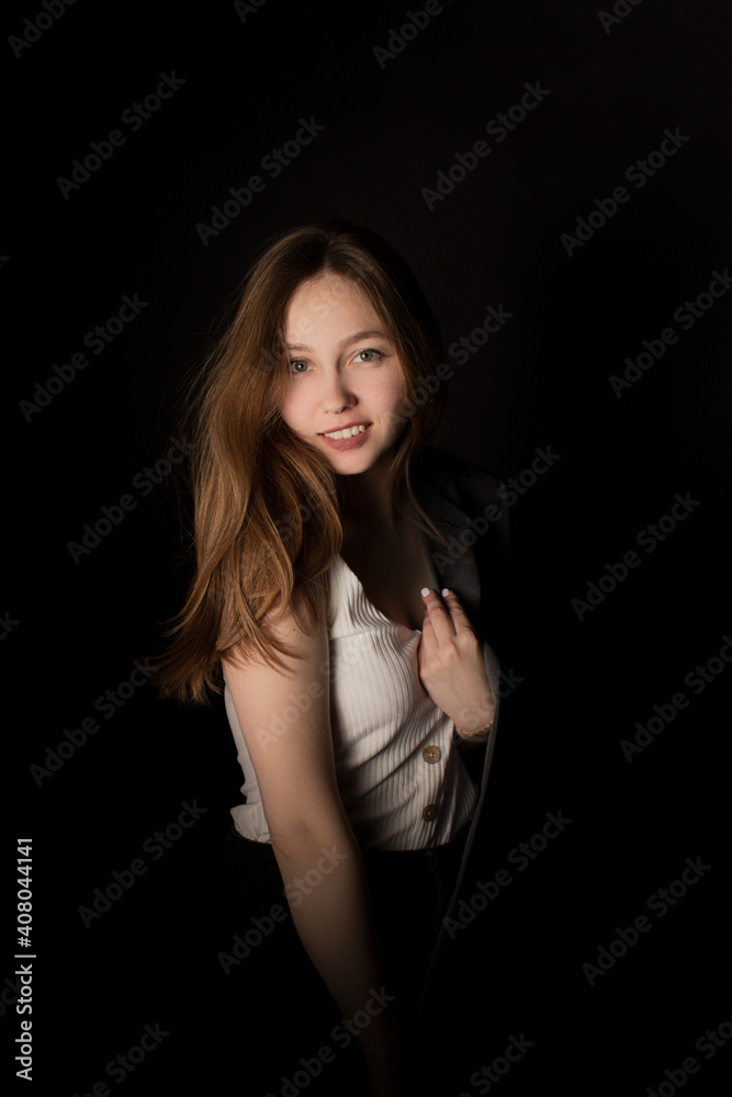 Young woman in jacket and white t-shirt standing alone in dark room and looking at camera, black background. Studio shot in low key. Casual portrait of girl with natural makeup and clean skin. Young woman in jacket and white t-shirt standing alone in dark room and looking at camera, black background. Studio shot in low key. Casual portrait of girl with natural makeup and clean skin.